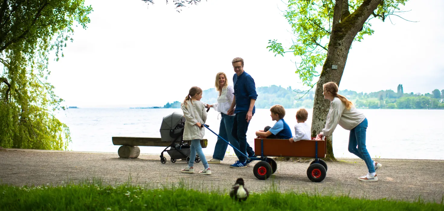 Familie mit zwei Erwachsenen und vier Kindern zieht einen roten Bollerwagen am Seeufer unter einem Baum