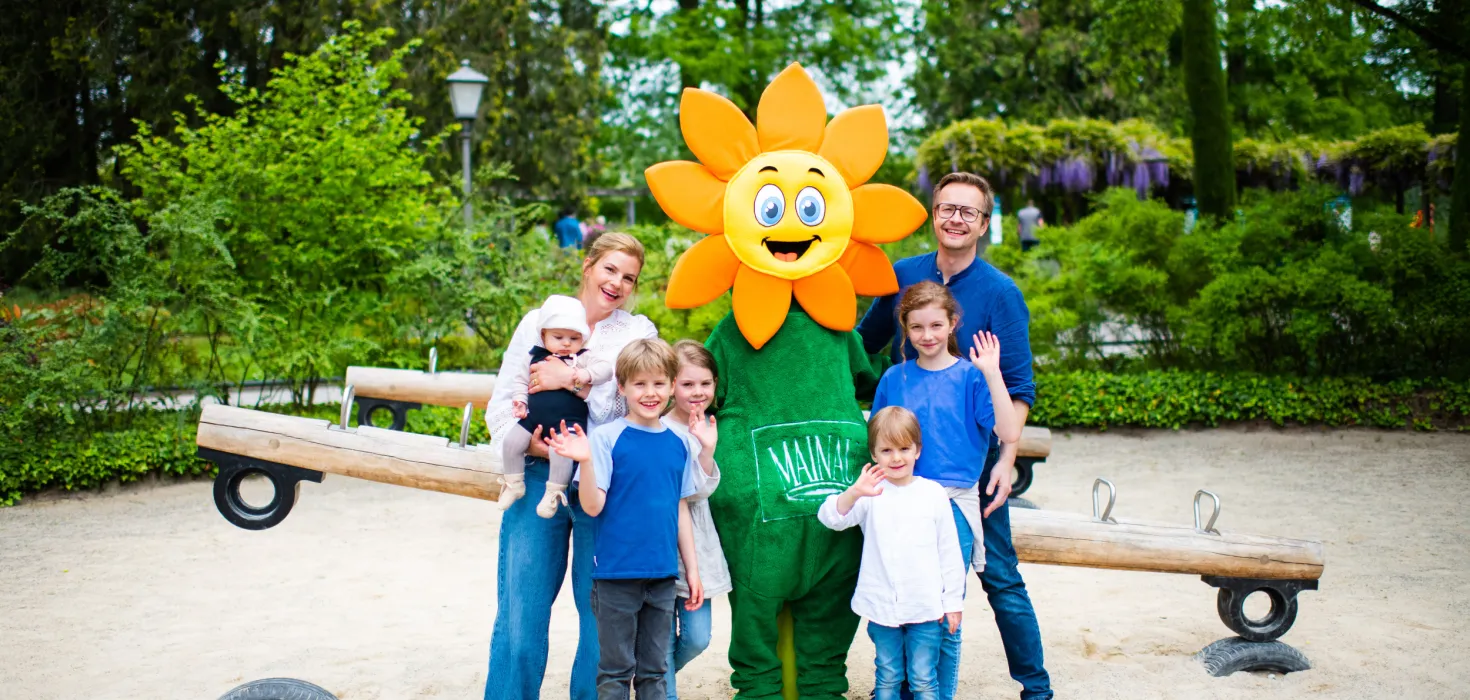 Gruppe von Kindern und Erwachsenen steht um das Maskottchen Blumi auf einem Spielplatz mit Wippe und Bäumen im Hintergrund
