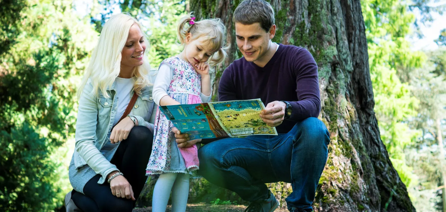 Familie mit zwei Erwachsenen und einem Kind kniet auf Gras vor einem Baumstamm und betrachtet gemeinsam eine Schatzkarte