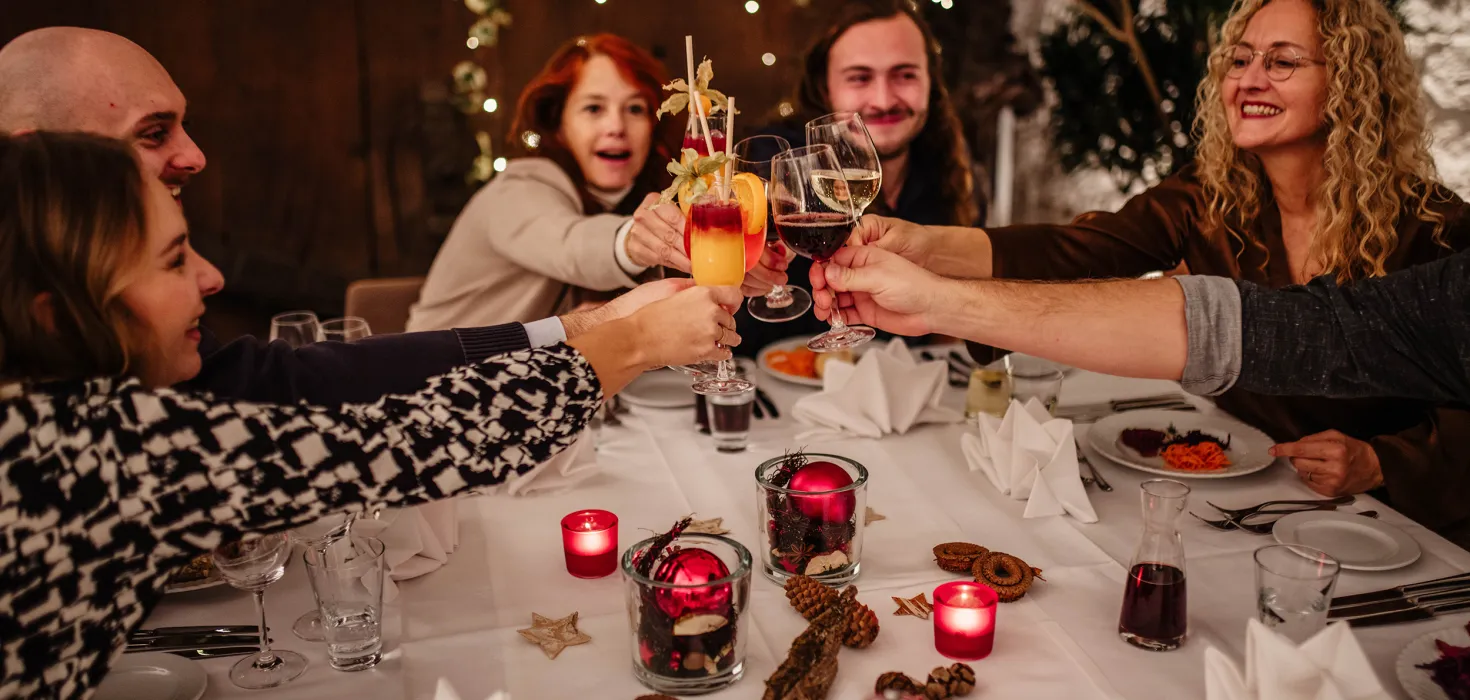 Eine Gruppe lachender Menschen sitzt an einem weihnachtlich dekorierten Tisch und stößt feierlich mit Getränken an