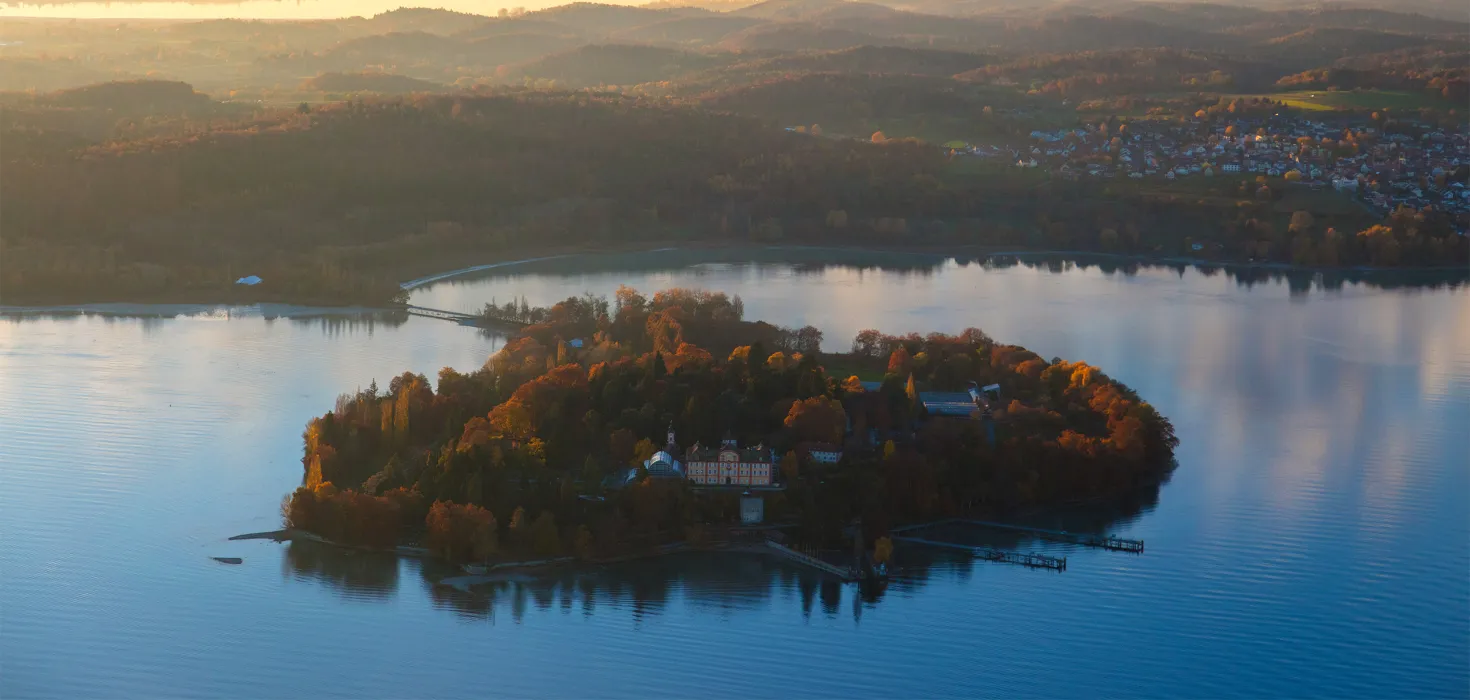 Luftaufnahme von der Insel Mainau im Herbst. Im Hintergrund ist die umgebende Landschaft sowie Nebelfelder zu sehen sowie die Herbstfärbung der Bäume