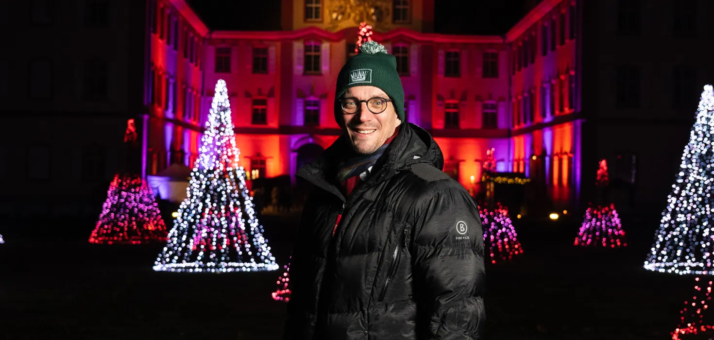 Ein Mann mit warmer Kleidung steht vor einem beleuchteten Gebäude und lacht in die Kamera