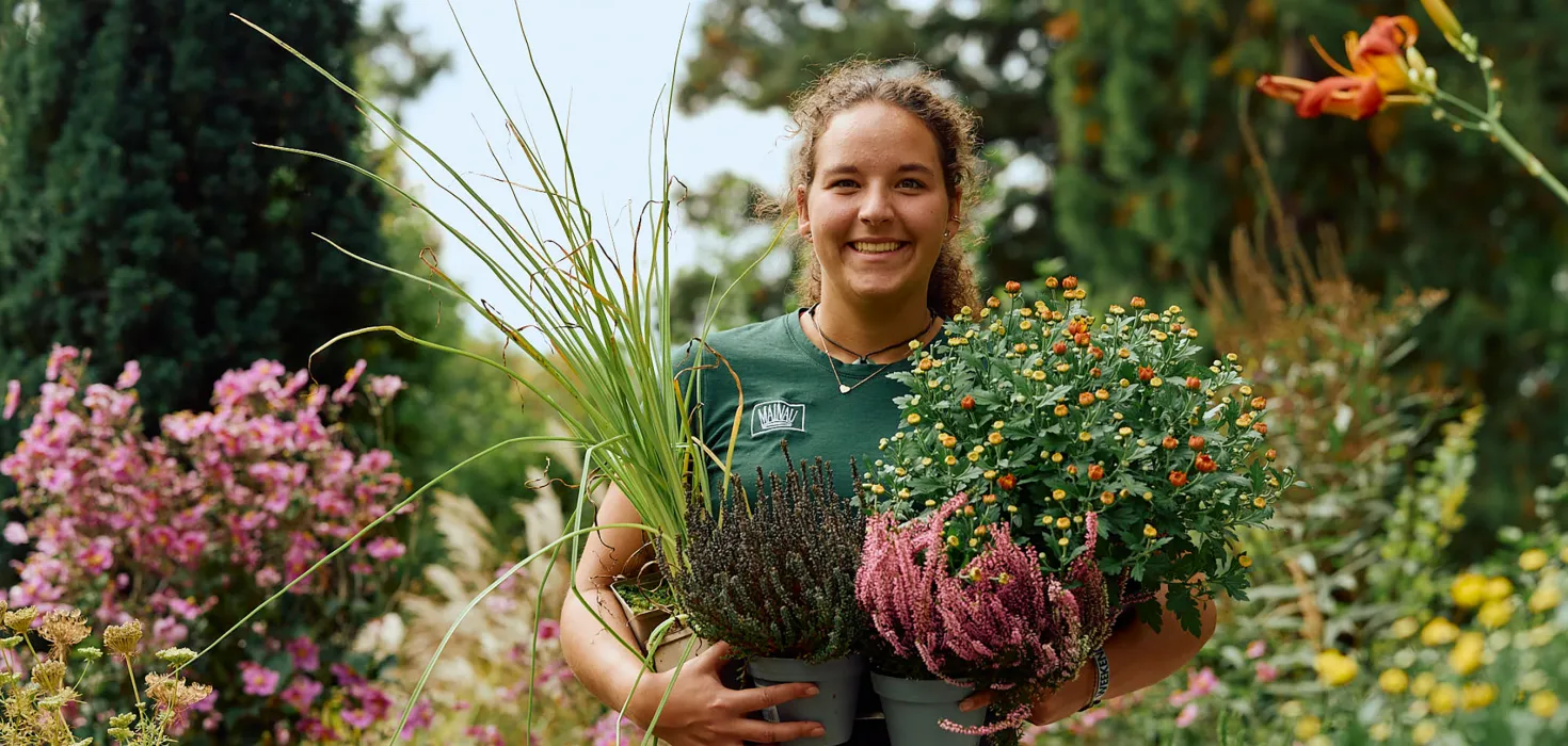 Eine junge, lächelnde Frau, die eine Schale voller bunter Blumen und Pflanzen hält, steht in einem blühenden Garten.