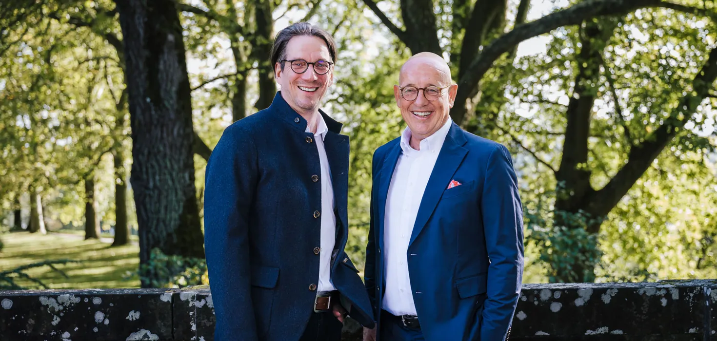 Zwei lächelnde Männer stehen in einem Park vor grünen Bäumen.