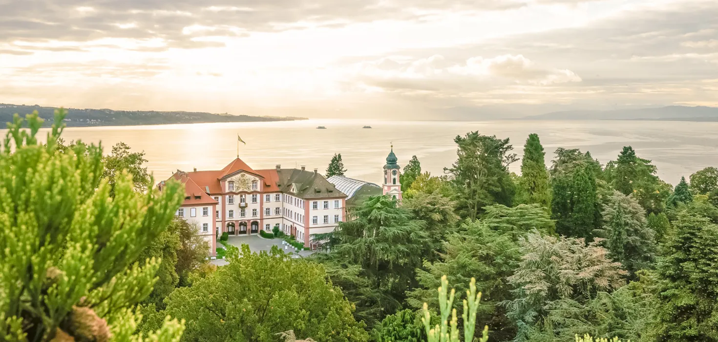 Blick über eine grüne Parklandschaft auf ein historisches Gebäude am Ufer eines Sees, im Hintergrund ruhiges Wasser und bewölkter Himmel im Abendlicht.