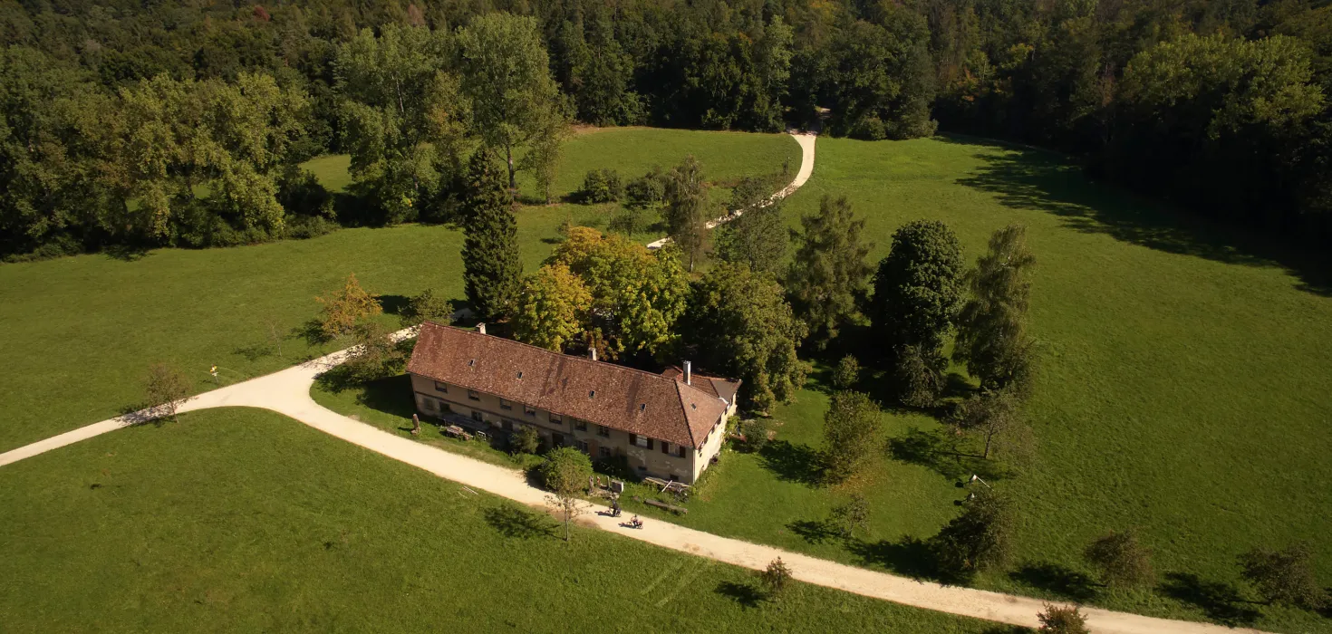 Luftaufnahme eines Hauses inmitten grüner Wiesen und Bäume, mit geschwungenem Weg und Wald, im Hintergrund ein See