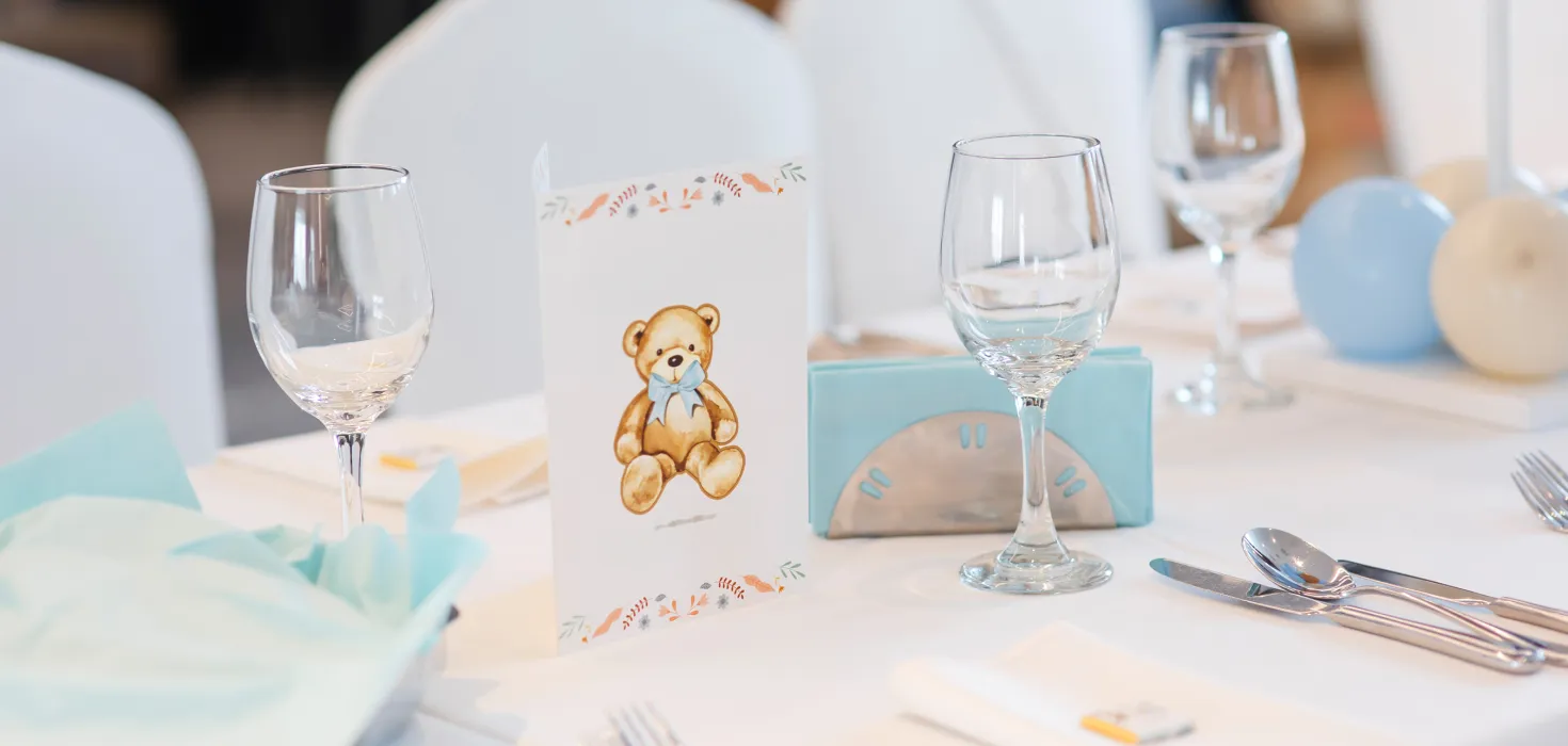 Festlich gedeckter Tisch mit Gläsern und Besteck, in der Mitte eine Tischkarte mit einem Teddybärenmotiv und hellblauer Dekoration