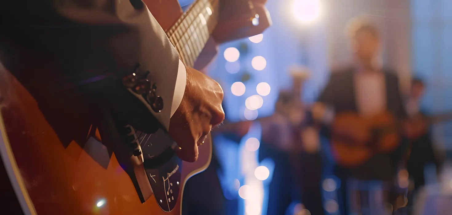 Nahaufnahme einer Person, die eine Akustikgitarre spielt, im Hintergrund sorgen Musiker und warmes Lichterambiente für eine stimmungsvolle Atmosphäre