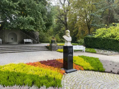 Büste auf schwarzem Sockel in einem parkähnlichen Bereich mit bunten Heiden Pflanzen und weißen Bänken
