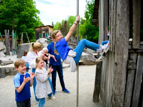 Kinder spielen auf dem Mainau Kinderland Spielplatz, ein Mädchen klettert an einem Pfosten hoch, andere Kinder und Erwachsene beobachten es