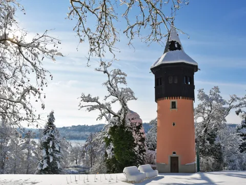 Schlanker runder Turm mit spitzem Dach in verschneiter Winterlandschaft und kahlen Bäumen