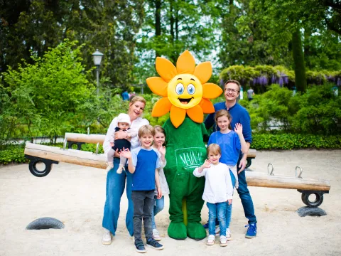 Gruppe von Kindern und Erwachsenen steht um das Maskottchen Blumi auf einem Spielplatz mit Wippe und Bäumen im Hintergrund