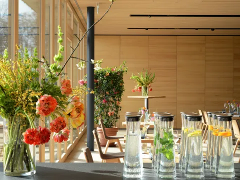 Glasvasen mit bunten Blumen und Karaffen mit Wasser und Früchten auf einem Tisch in einem hellen, modernen Raum mit Holzwänden und großen Fenstern