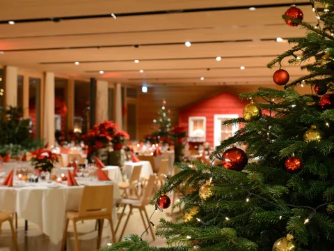 Weihnachtsbaum mit Kugeln im Vordergrund und festlich gedeckte Tische in einem Saal im Hintergrund