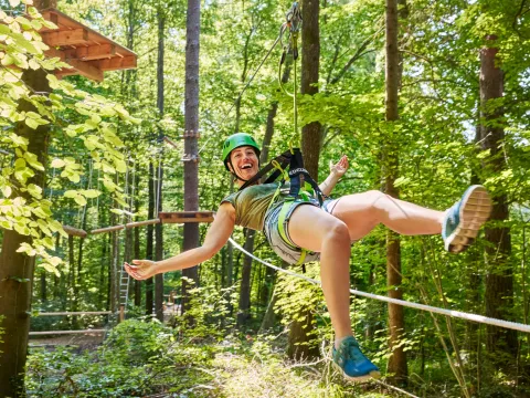 Mainau Erlebniswald / Anna Glad Junge Frau hängt fröhlich an einem Kletterseil und freut sich