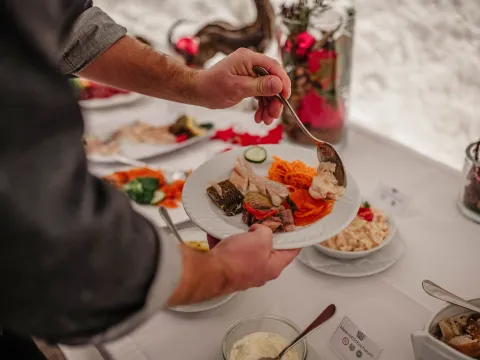 Schwedisches Weihnachtsbuffet Person, die sich an einem Buffet den Teller füllt und mit einem Löffel Speisen auf einen weißen Teller gibt, auf dem bereits eingelegtes Gemüse, geraspelte Karotten, Gurkenscheiben, marinierter Fisch und Aufschnitt liegen. Der Buffettisch ist mit Schüsseln mit Salaten, Saucen und Brot gedeckt und mit roten Weihnachtsornamenten und Kerzen dekoriert, um eine festliche Atmosphäre zu schaffen.