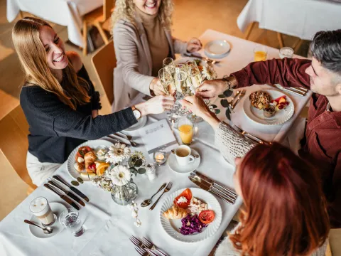 Sonntagsbrunch Eine Gruppe von Freunden genießt einen Brunch in einem Restaurant und erhebt ihre Champagnergläser zu einem Toast über einer weißen Tischdecke. Es werden Croissants, frisches Obst, Tomaten, Rotkohlsalat, Müsli mit Joghurt und Räucherlachs serviert, dazu Kaffee, Orangensaft und Latte Macchiato. Der Tisch ist mit einer Vase mit weißen Blumen dekoriert, was eine festliche und elegante Atmosphäre schafft