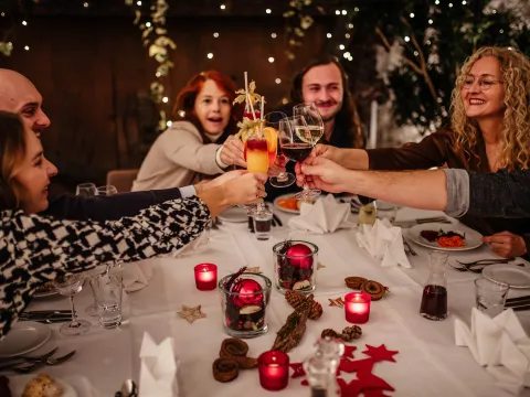 Weihnachtsmenü mit Familie & Freunde Eine Gruppe lachender Menschen sitzt an einem weihnachtlich dekorierten Tisch und stößt feierlich mit Getränken an