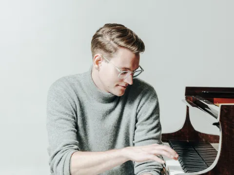 Mann mit blonden Haaren sitzt an einem Klavier, trägt einen grauen Pullover und schwarze Hosen, spielt mit der rechten Hand auf den Tasten.