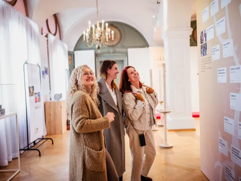 Ausstellung im Schloss Mainau Drei lächelnde Frauen betrachten eine Ausstellung in einem hell erleuchteten Raum. Die Frauen tragen legere Mäntel und Jacken und stehen gemeinsam vor einer großen Wandtafel mit Namen und Text, wahrscheinlich einem Stammbaum oder einer historischen Zeitleiste. Der Raum ist elegant eingerichtet mit hohen Gewölbedecken, Kronleuchtern, weißen Vorhängen und Vitrinen, was eine raffinierte und kulturelle Atmosphäre schafft. Die Frauen wirken interessiert und fasziniert von der Ausstellung.