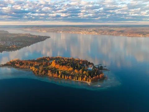 Luftaufnahme von der Insel Mainau im Herbst. Im Hintergrund ist die umgebende Landschaft sowie Nebelfelder zu sehen sowie die Herbstfärbung der Bäume