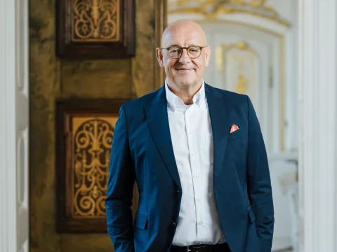 Ein lächelnder Mann im Anzug steht vor einem geöffneten Saal