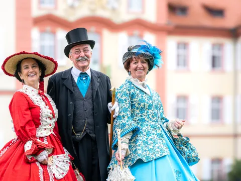 Gräfliches Schlossfest Zwei Frauen und ein Mann in historischen Kostümen lachen in die Kamera und stehen vor einem schlossähnlichem Gebäude