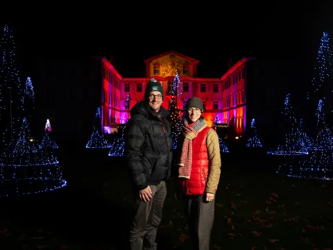 Ein Mann und eine Frau in warmer Kleidung stehen vor einem Gebäude, das angestrahlt wird. Neben ihnen sind Lichterketten in Form von Bäumen zu sehen.