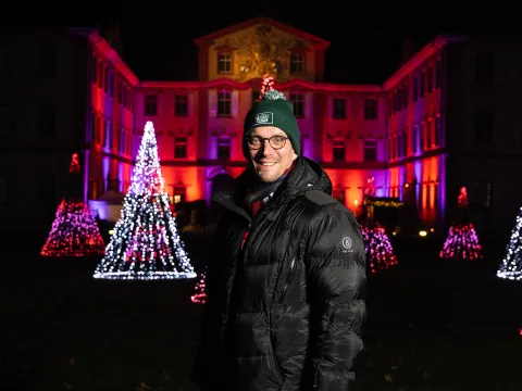 Ein Mann mit warmer Kleidung steht vor einem beleuchteten Gebäude und lacht in die Kamera