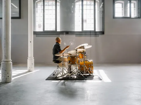 Mann sitzt an einem Schlagzeug auf einem Teppich in einem großen, hellen Raum mit hohen Fenstern und Säulen.
