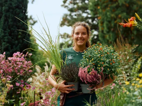 Eine junge, lächelnde Frau, die eine Schale voller bunter Blumen und Pflanzen hält, steht in einem blühenden Garten.