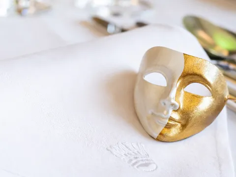 Eine goldene, dekorative Theatermask liegt auf einer weißen Stoffserviette, im Hintergrund sind Gläser eingedeckt