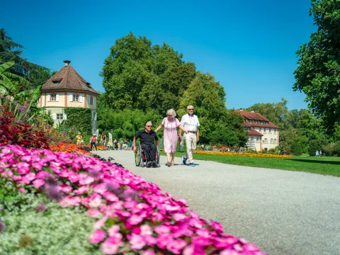 Drei ältere Menschen spazieren gemeinsam durch einen Park, eine Person im Rollstuhl, umgeben von bunten Blumenbeeten und historischen Gebäuden