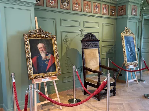Ausstellungsraum zur Ausstellung „200 Jahre Großherzog Friedrich“ mit einem historischen Holzstuhl, zwei gerahmten Porträts auf Staffeleien und rot abgesperrtem Bereich vor grüner Wandvertäfelung. An der oberen Wand sind mehrere Wappen und Namensschilder angebracht.