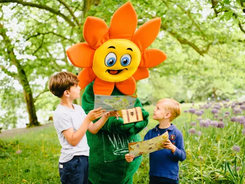 Zwei Kinder stehen mit einer blumenförmigen Maskottchenfigur im Park der Insel Mainau und betrachten eine Schatzkarte sowie eine kleine Truhe. Die Szene zeigt eine Kinderschatzsuche in einer Gartenlandschaft mit Bäumen und Blumen im Hintergrund.