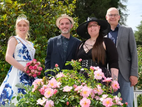 Vier Personen stehen bei einer Rosentaufe in einem Garten vor einem blühenden Rosenbeet; links eine junge Frau mit Krone, Schärpe und Rosenstrauß, daneben drei weitere Personen. Im Vordergrund sind rosa Rosen zu sehen, im Hintergrund Bäume und Sträucher.