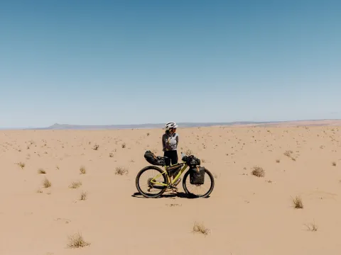 Eine Radreisende steht mit ihrem voll bepackten Fahrrad in einer weitläufigen Sandwüste in Namibia. Im Hintergrund sind vereinzelte Sträucher und eine flache Berglandschaft zu sehen.