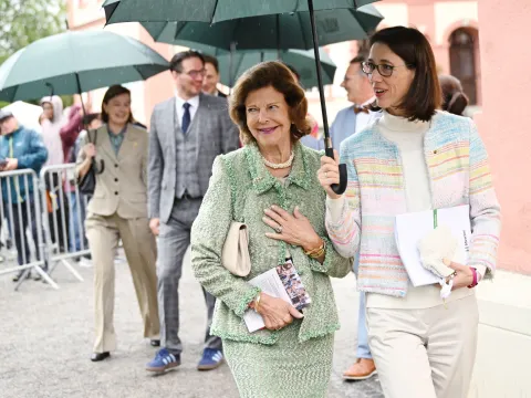 Königin Silvia von Schweden geht bei ihrem Besuch auf der Insel Mainau im Regen neben Bettina Gräfin Bernadotte, die einen Regenschirm über sie hält. Im Hintergrund sind weitere Begleitpersonen und wartende Menschen zu sehen.