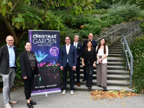 Sieben Personen stehen bei einem Pressetermin zum Christmas Garden Insel Mainau neben einem Veranstaltungsplakat vor einer Außentreppe. Das Plakat kündigt den Christmas Garden vom 19. November 2026 bis zum 9. Januar 2027 an.