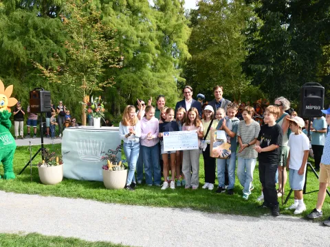 Eine Gruppe von Schülerinnen und Schülern präsentiert bei der Prämierung der Vogelscheuchen-Ausstellung „Seegestalten“ auf der Insel Mainau einen Scheck für den ersten Platz. Hinter der Gruppe stehen zwei Erwachsene, links ist das Mainau-Blumenmaskottchen zu sehen.
