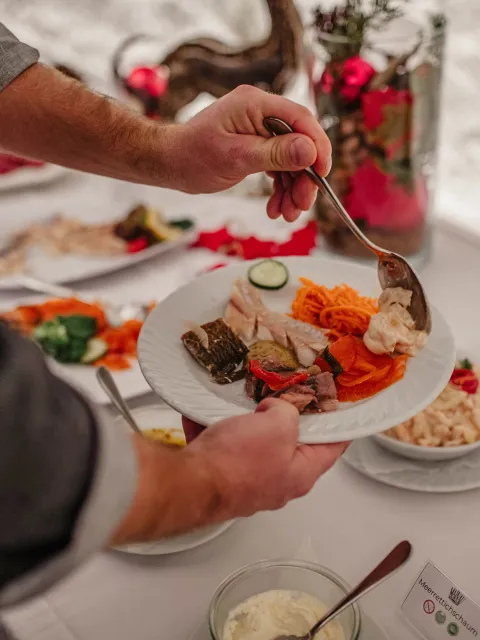 Person, die sich an einem Buffet den Teller füllt und mit einem Löffel Speisen auf einen weißen Teller gibt, auf dem bereits eingelegtes Gemüse, geraspelte Karotten, Gurkenscheiben, marinierter Fisch und Aufschnitt liegen. Der Buffettisch ist mit Schüsseln mit Salaten, Saucen und Brot gedeckt und mit roten Weihnachtsornamenten und Kerzen dekoriert, um eine festliche Atmosphäre zu schaffen.