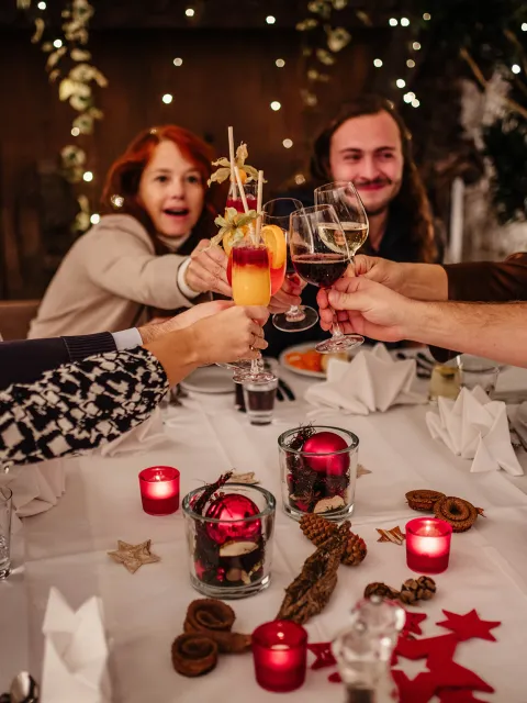 Eine Gruppe lachender Menschen sitzt an einem weihnachtlich dekorierten Tisch und stößt feierlich mit Getränken an