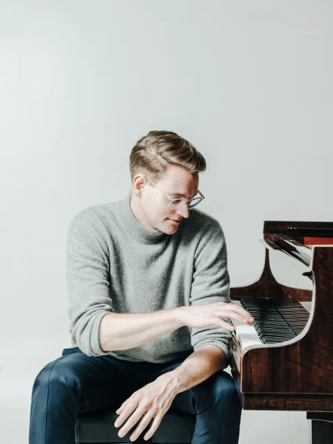 Steinway Artist Maximilian Schairer Mann mit blonden Haaren sitzt an einem Klavier, trägt einen grauen Pullover und schwarze Hosen, spielt mit der rechten Hand auf den Tasten.