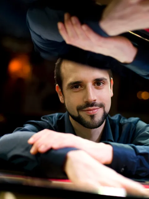 Männliche Person lehnt mit verschränkten Armen auf einem Klavier, im Hintergrund unscharfe Lichter und lächelt in die Kamera