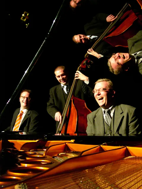 Drei Musiker in Anzügen spielen Klavier, Kontrabass und Schlagzeug in dunkler Umgebung.