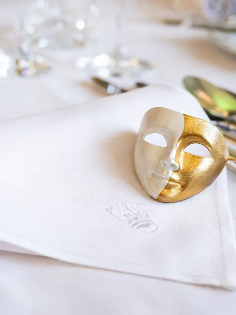 Eine goldene, dekorative Theatermask liegt auf einer weißen Stoffserviette, im Hintergrund sind Gläser eingedeckt