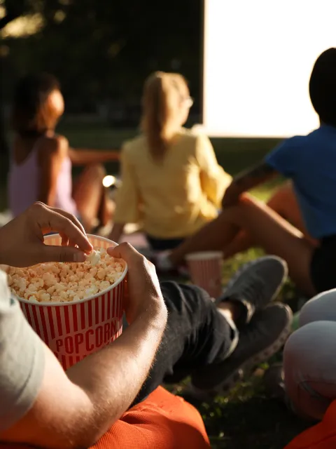 Menschen sitzen auf Sitzsäcken auf einer Wiese und schauen gemeinsam ein Open-Air-Kino, während eine Person Popcorn aus einem Becher isst.