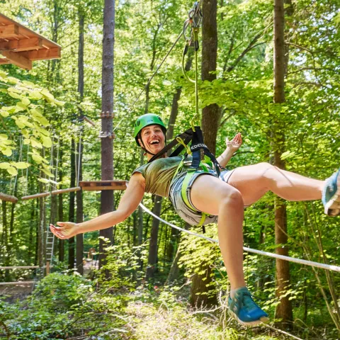 Mainau Erlebniswald / Anna Glad Junge Frau hängt fröhlich an einem Kletterseil und freut sich