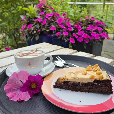 Kaffee und Kuchen im Café Vergissmeinnicht Ein Stück Schokoladenkuchen liegt auf einem Teller mit rotem Porzellanrand. Der Teller befindet sich auf einem schwarzen Tablett, ebenfalls darauf befindet sich eine Tasse Kaffee sowie eine rosa Blüte als Dekoration. Im Hintergrund sind bepflanzte Kübel und grüne Pflanzen zu sehen