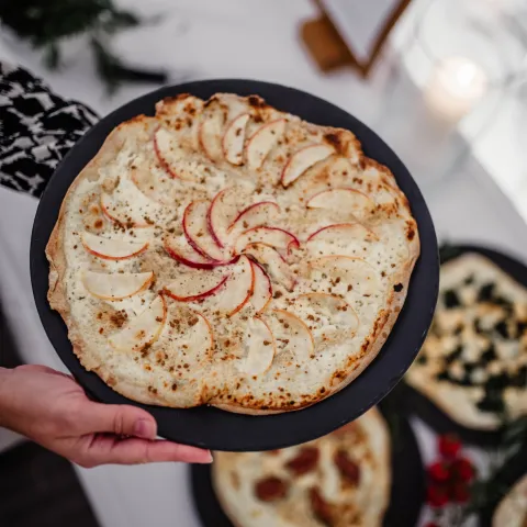 Dinnele Essen beim Christmas Garden Insel Mainau Ein mit Apfel belegter Hefeteig auf einem Teller, der von zwei Händen gehalten wird. Im Hintergrund sind weitere Dinnele abgebildet